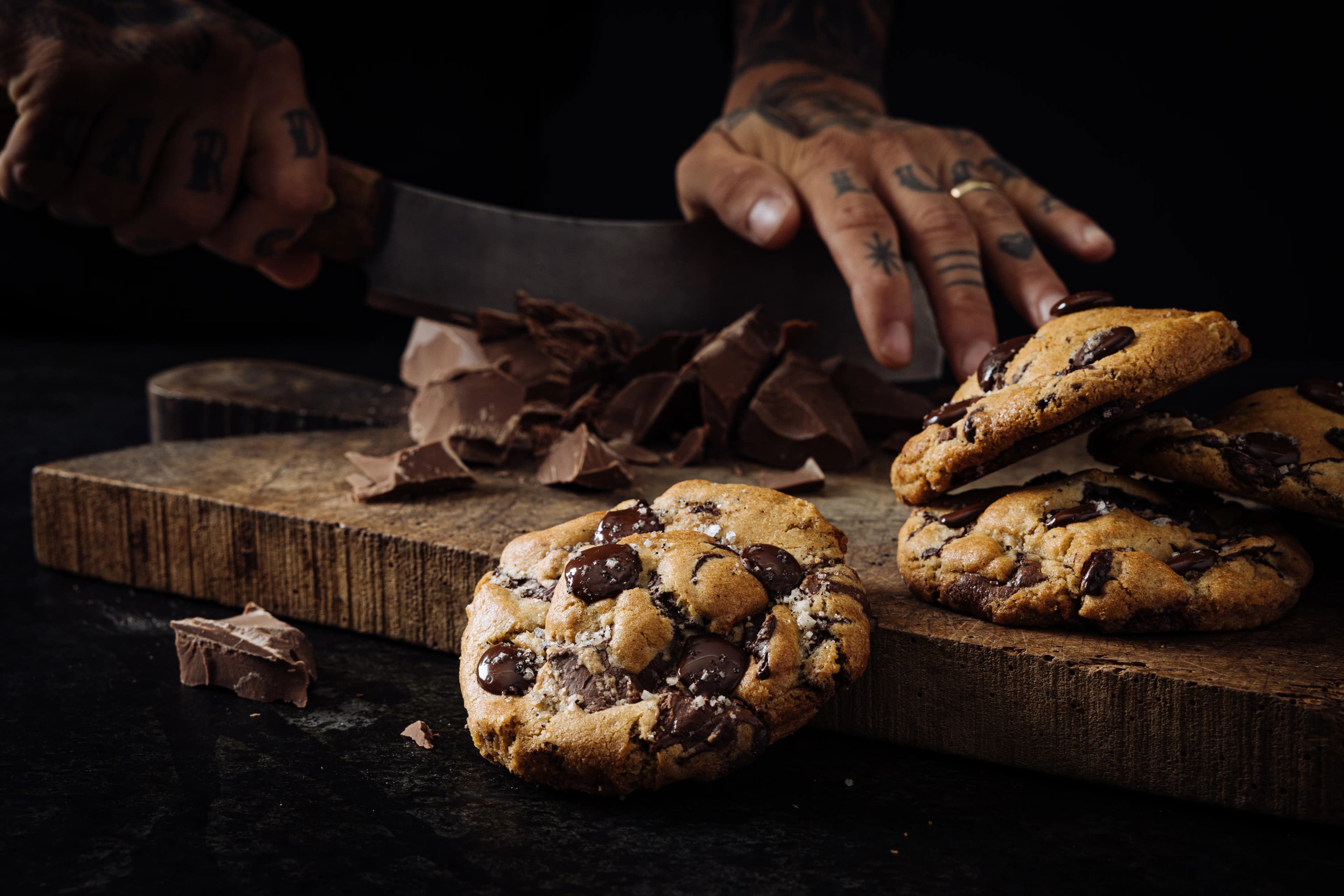 Hands chopping chocolate beside a finished chocolate chip cookie